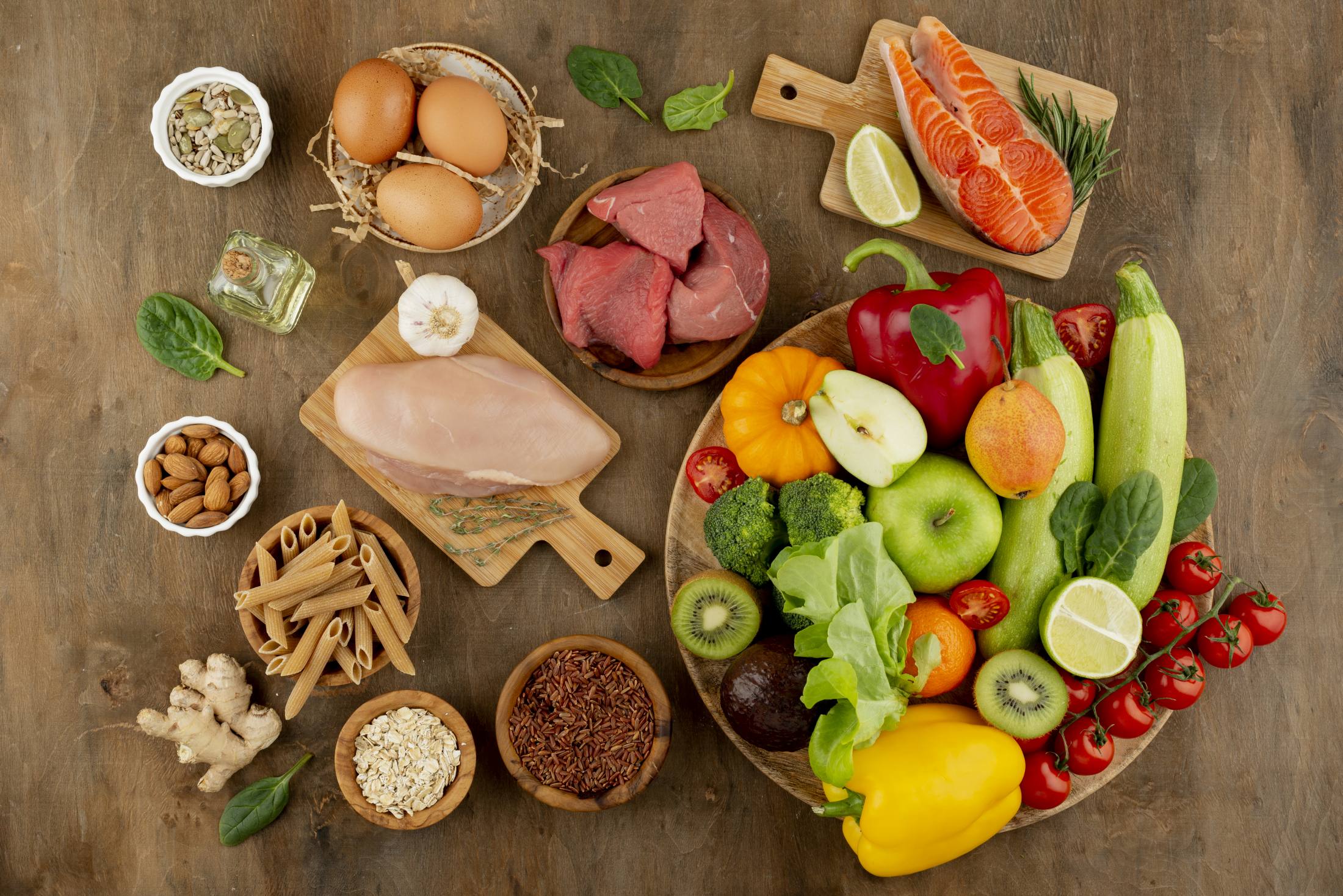 Różnorodne produkty spożywcze ułożone na drewnianym blacie, w tym warzywa, owoce, mięso, jaja, orzechy, makarony i olej